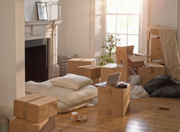 A room filled with unpacked moving boxes, cushions on the floor, a laptop on a box, and a window letting in sunlight