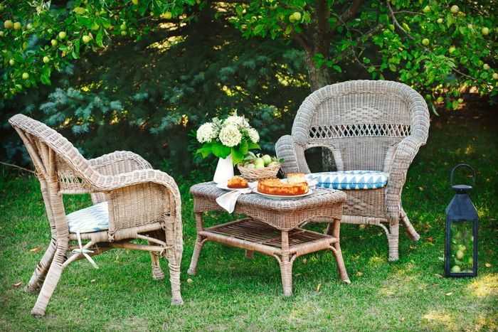 Two wicker chairs with cushions surrounding a wicker table adorned with flowers, a pie, and fresh fruit, placed on a lush lawn beneath green trees. A lantern with fruit rests nearby.