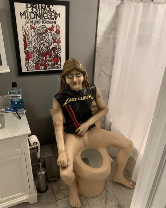 A sculpture of a person wearing a Lars Ulrich shirt, sitting on a toilet, placed next to a shower in a bathroom