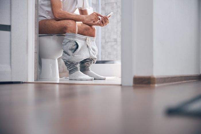 A person sitting on a toilet with their pants down, looking at a smartphone. The setting appears to be a modern bathroom design