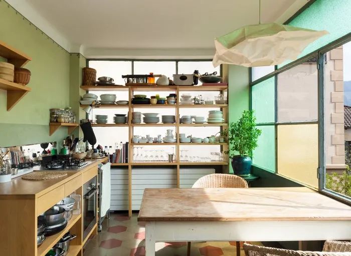 A well-organized kitchen featuring open shelving, an array of utensils, a central dining table, and lush greenery by the window.