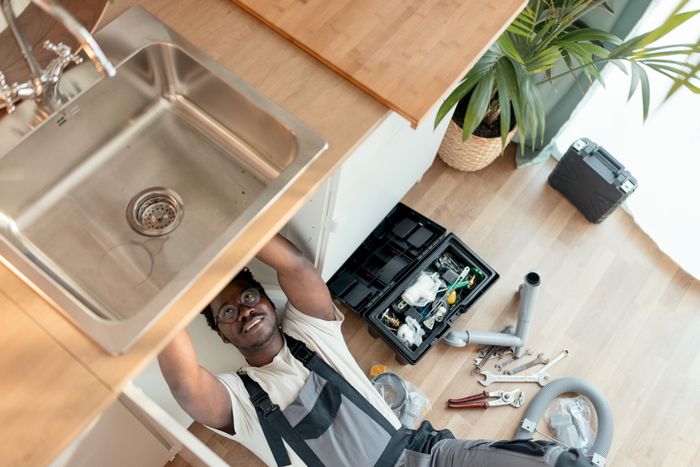 A plumber is lying on his back, repairing a sink. Tools and equipment are scattered on the floor beside him, along with a plant and a toolbox nearby