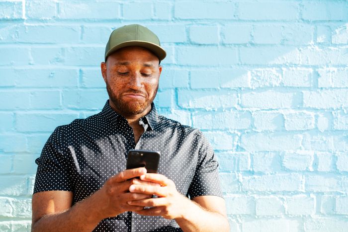 Man wearing a cap and a dotted shirt using a smartphone against a blue brick wall
