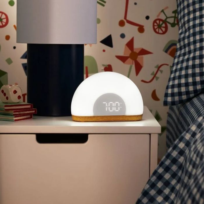 A modern digital alarm clock on a nightstand, showing 7:00 AM, with books and a lamp nearby, capturing a cozy home decor vibe.