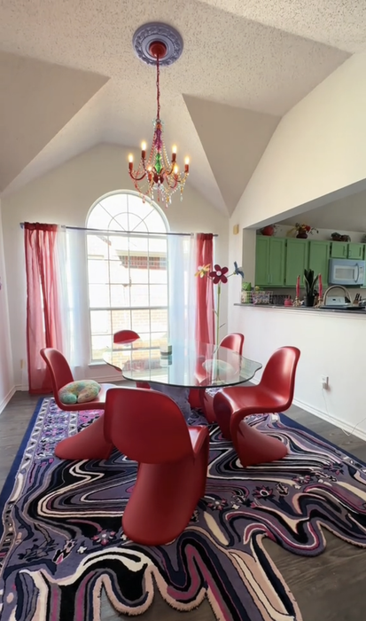 A modern interior room showcasing a glass dining table, red chairs, a colorful rug, and a statement chandelier