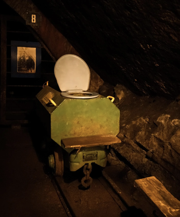 An old-fashioned wooden toilet cart with a hinged lid, placed in a dimly lit, rustic underground area
