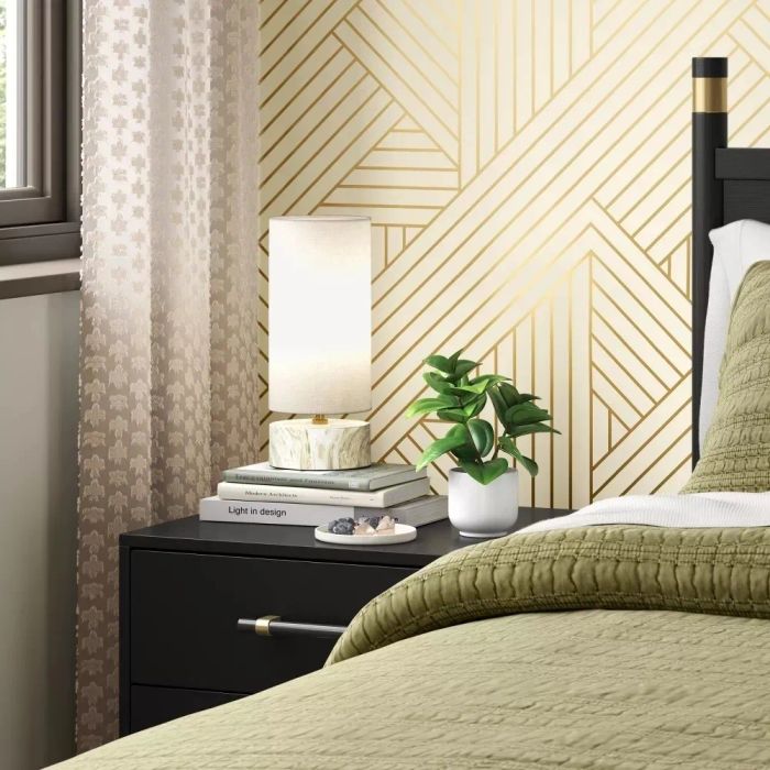 A contemporary bedroom featuring patterned wallpaper, a bedside table adorned with a lamp, books, a plant, and a cozy, textured bedspread.