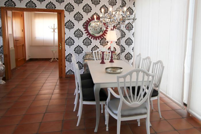 A chic dining room featuring a white table with six matching white chairs, an elegant wall decor, and a chandelier. In the background, you can see an adjacent room with tiled flooring.