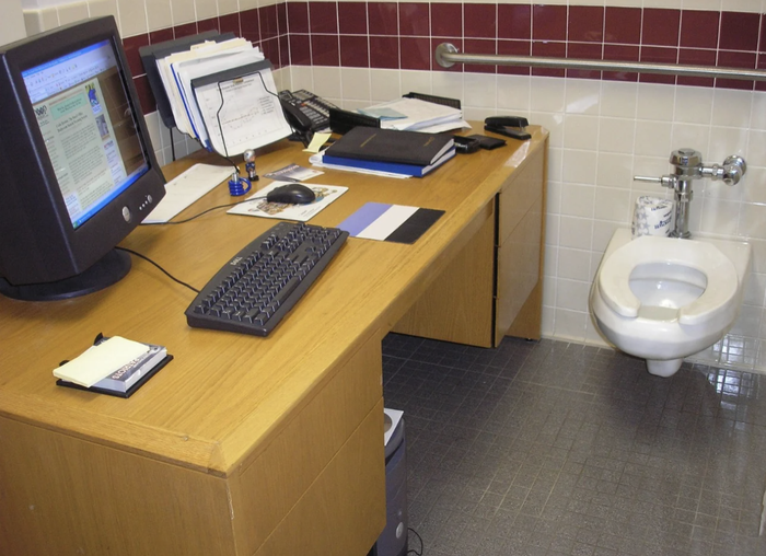 An office desk with a computer setup placed in a bathroom beside a toilet