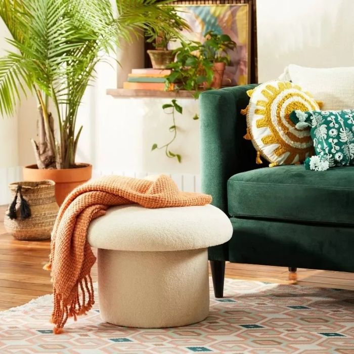 A cozy living space featuring a green sofa adorned with decorative cushions, a small white stool draped with an orange blanket, and lush potted plants surrounding the room.