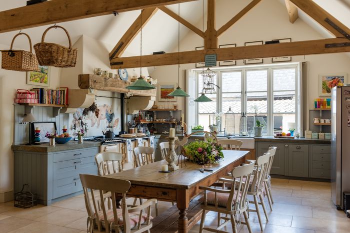 A spacious rustic kitchen with exposed wooden beams, a large dining table surrounded by chairs, hanging light fixtures, and an assortment of shelves, cabinets, and decorative accents.