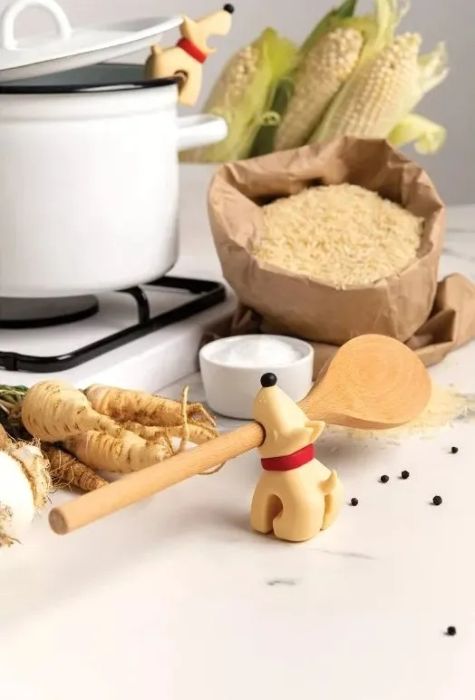 Dog-shaped spoon rest with a wooden spoon on a kitchen counter, surrounded by a pot, corn, rice, and spices