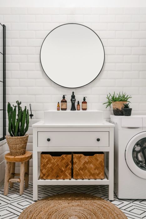 A contemporary bathroom featuring a round mirror, elegant sink vanity, woven storage baskets, and a washing machine placed beside a vibrant potted plant.