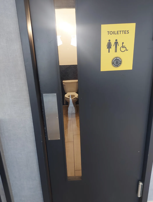 A public restroom door featuring a window that reveals the toilet to anyone standing outside