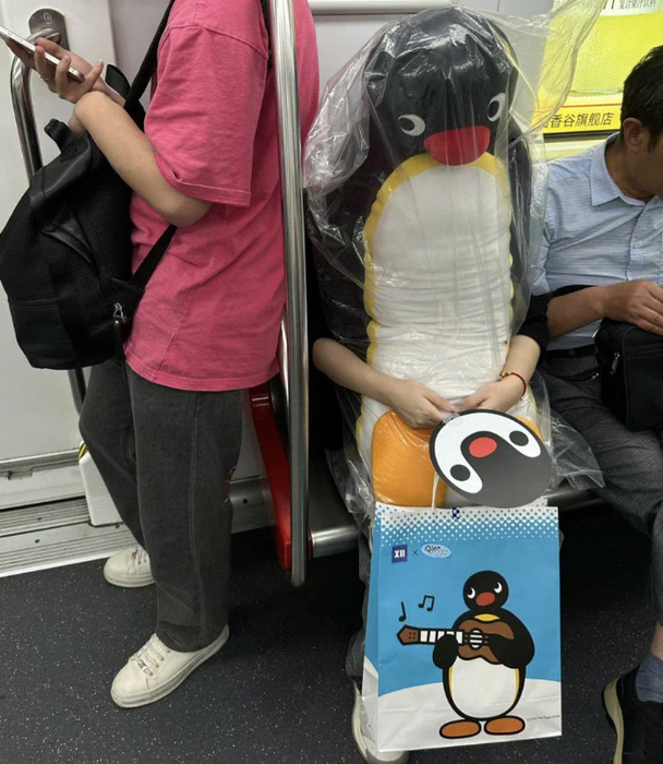 A person seated on a subway train, clad in a Pingu outfit topped with a plastic raincoat and holding a matching bag. Another passenger stands nearby, dressed in everyday attire.