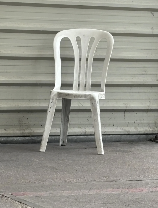 A white plastic chair positioned in front of a corrugated metal wall, resting on a concrete surface.