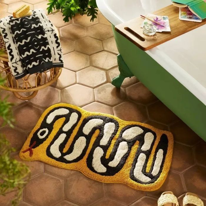 A snake-shaped bath mat resting on a tiled bathroom floor, accompanied by a green bathtub and a basket full of towels.