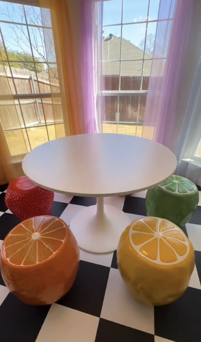 A table surrounded by fruit-shaped stool chairs on a checkered floor, with purple curtains framing the window