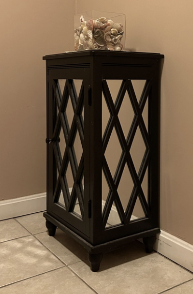 A black cabinet with a glass door adorned with diamond-shaped designs, positioned on a tiled floor. A glass jar filled with seashells sits on top.