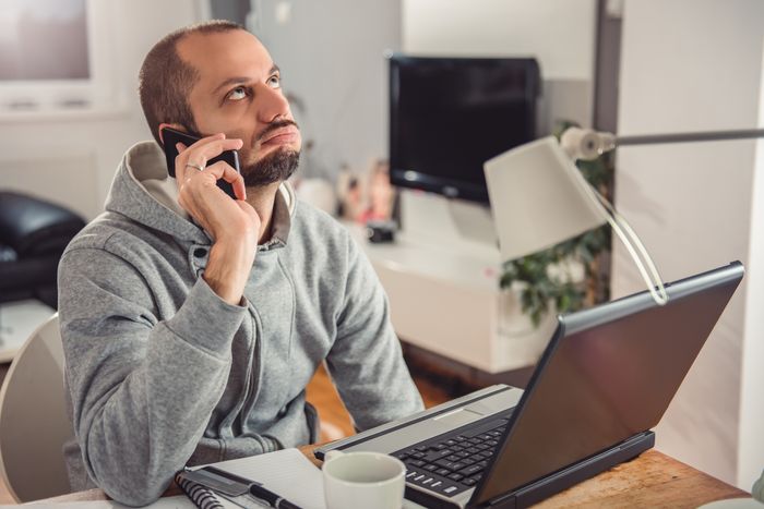 Person in a hoodie sitting at a desk with a laptop, speaking on the phone