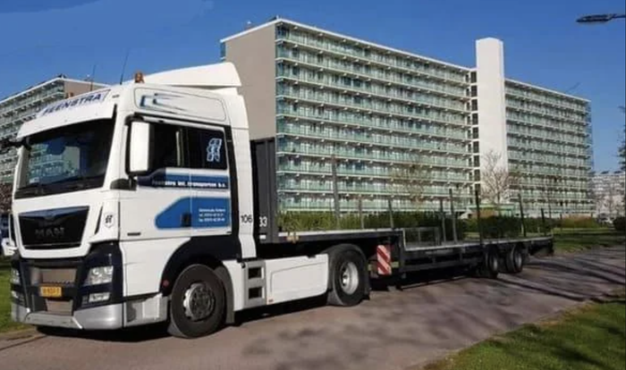 A sizable truck equipped with a flatbed trailer stationed in a neighborhood, with an apartment complex visible behind it.