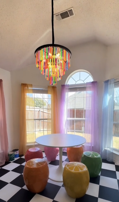 A view of the breakfast nook: A colorful spoon chandelier hangs above a white table surrounded by multicolored fruit chairs, with curtains in different colors hanging in the background