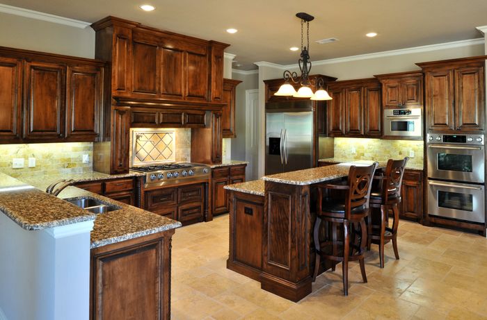 A spacious kitchen with wooden cabinets, granite countertops, stainless steel appliances, and an island featuring bar stools. Chandeliers are suspended from the ceiling.