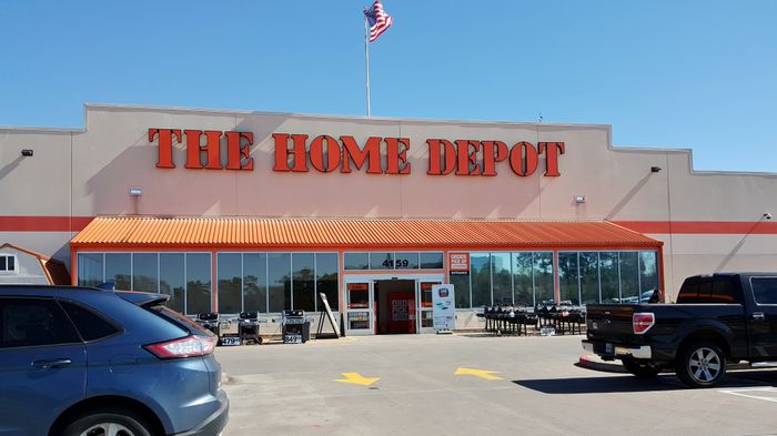 The Home Depot storefront with cars parked in front, American flag flying above, and the signature orange roof canopy visible