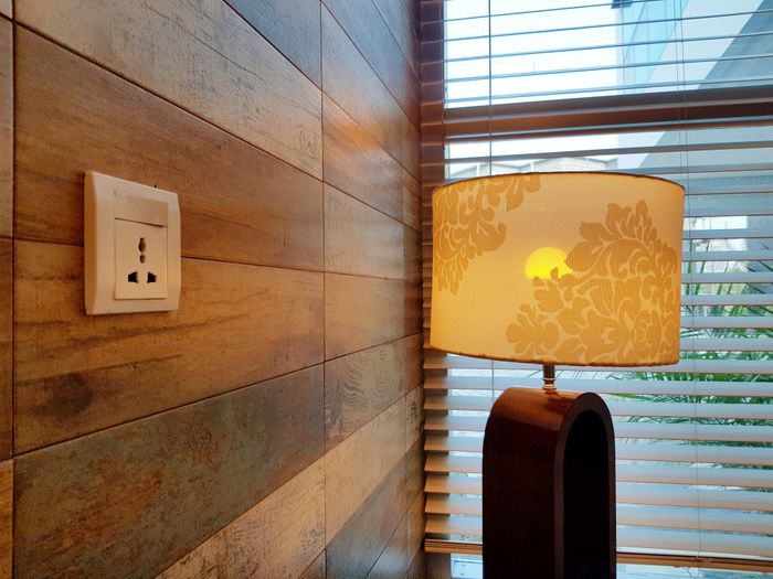A wooden wall with an electrical outlet beside a lamp topped with a floral-patterned shade, next to a window covered by blinds.
