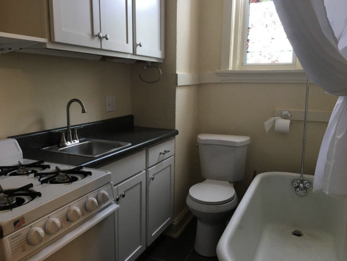 A small kitchen in an apartment with appliances next to an open bathroom that has a toilet and tub, showcasing a unique layout