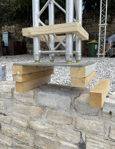 A temporary support structure is fashioned from wooden blocks and a metal sheet, propping up a metal frame against a stone wall. The scene is devoid of any people.
