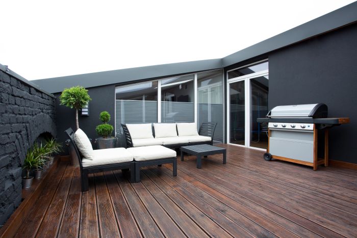 A stylish outdoor patio adjacent to a house with a sleek black exterior, featuring a cozy seating area and a barbecue grill.