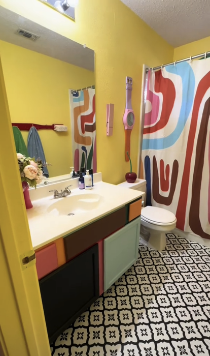 A vibrant bathroom featuring an abstract-patterned shower curtain, a multicolored cabinet, and a geometric-tiled floor, complete with a vanity filled with various toiletries