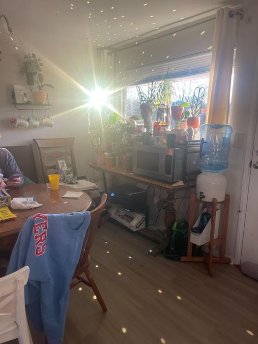 A kitchen featuring a table, chairs, a blue sweatshirt draped over a chair, and a water cooler. Sunlight streams through a window, casting dazzling light patterns across the room.