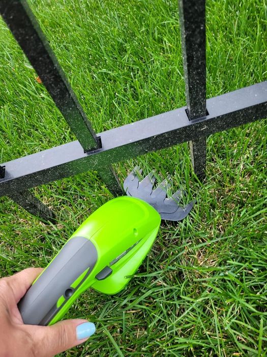 Hand holding a green gardening tool near a metal fence, with neatly trimmed grass visible