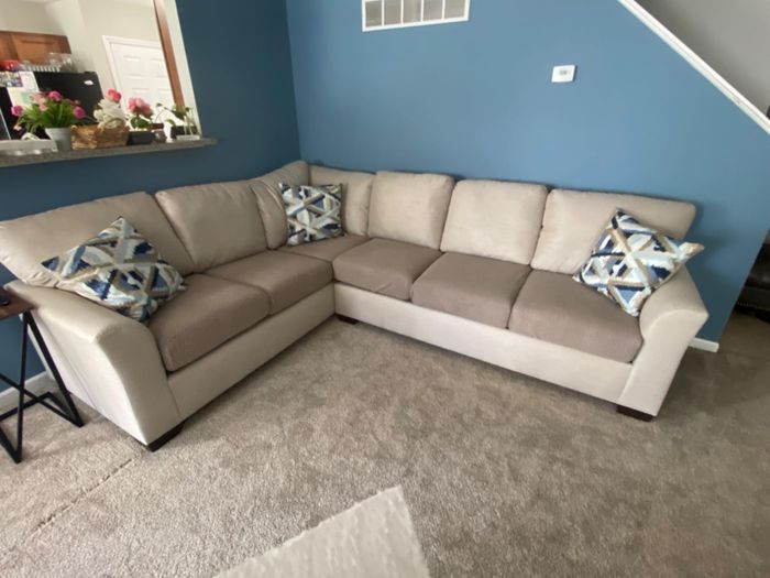 A beige L-shaped sectional sofa with a variety of patterned throw pillows, placed in a living room featuring blue walls and a light gray carpet.