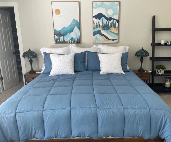 A beautifully arranged bed with a blue comforter, matching white and blue pillows, placed between two nightstands with decorative lamps, and adorned with wall art above
