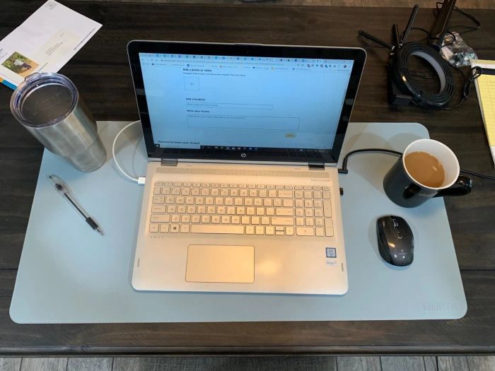 A reviewer's photo showing the rectangular desk mat in blue, with a laptop, coffee cup, mouse, and pen on top.