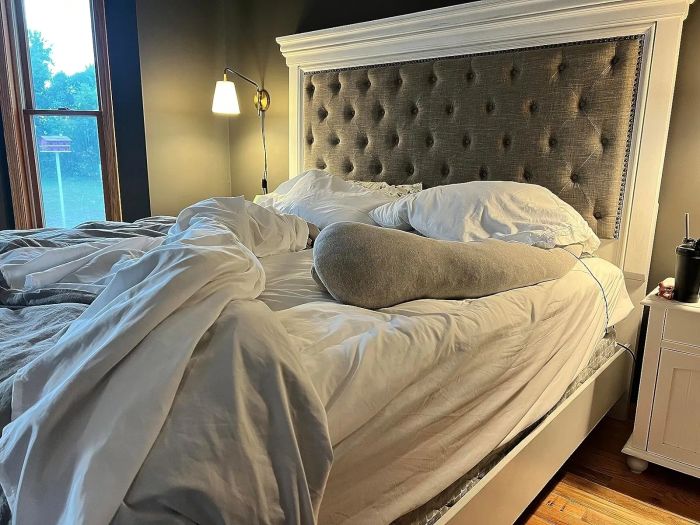 A disheveled bed featuring a large headboard and pillows, situated in a room with a nightstand and lamp