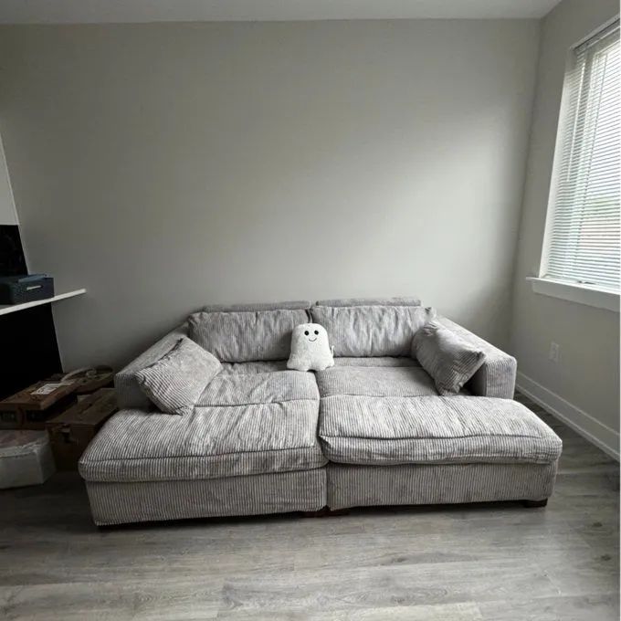 A stylish minimalist gray sofa adorned with an adorable ghost pillow, positioned against a plain wall with moving boxes nearby, bathed in soft natural light.