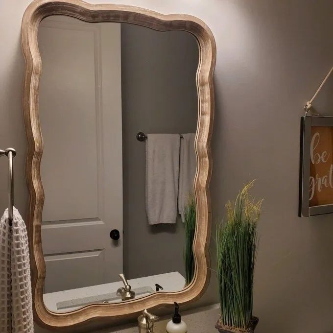 A bathroom mirror with a rustic wooden frame hanging above a sink, accompanied by a towel rack and a partially visible wall sign reading 'be gr,' likely hinting at a 'be grateful' message.
