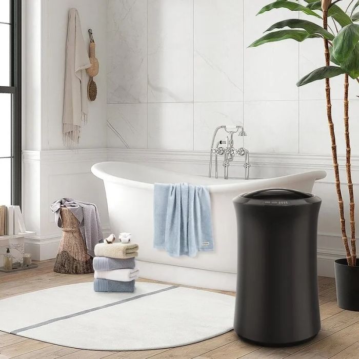 A sleek, modern bathroom featuring a freestanding tub, a chic black laundry bin, neatly stacked towels, and a lush green plant for a refreshing touch.