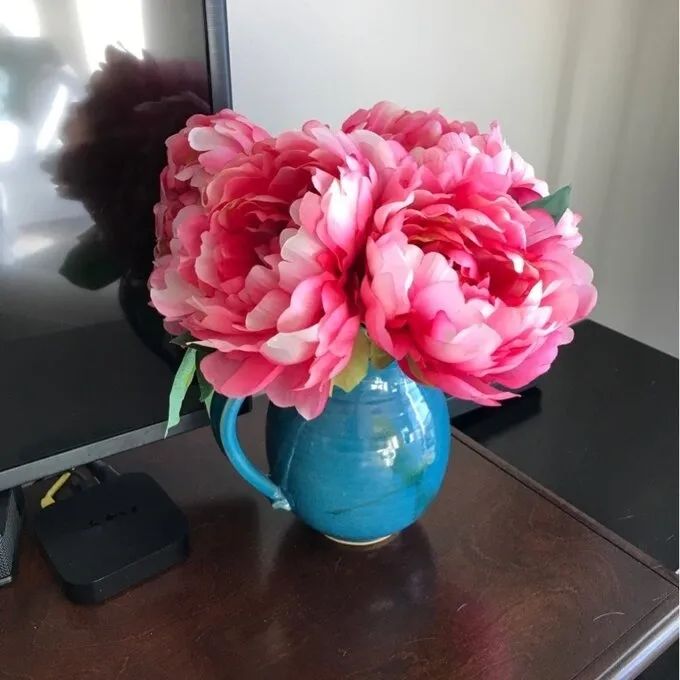 A delicate vase filled with pink peonies sits gracefully on a wooden table beside a television, adding a soft floral touch to the space.