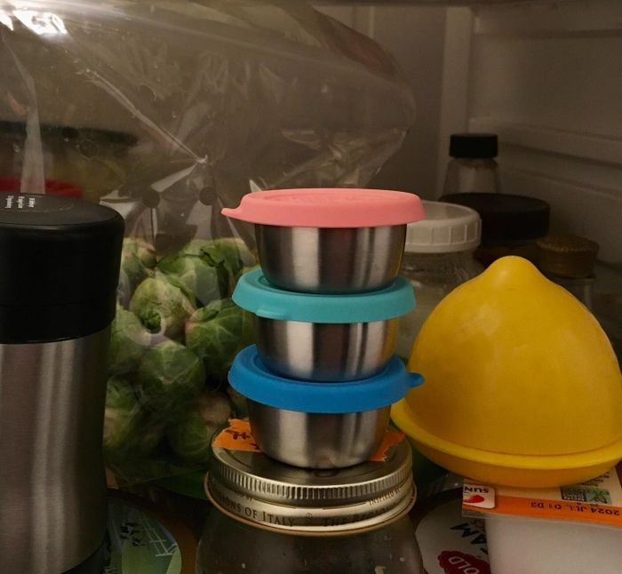 A stack of stainless steel condiment cups with colorful lids placed inside a refrigerator, surrounded by various jars and fresh produce