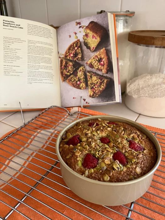 A freshly baked pistachio and raspberry cake cooling in a pan, with an open recipe book displaying the same cake in the background.