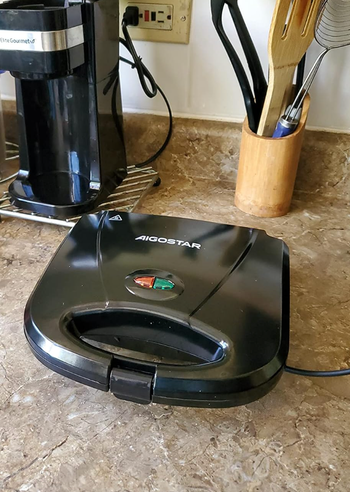 An Aigostar sandwich maker sitting on a kitchen counter, with utensils and a coffee maker in the background