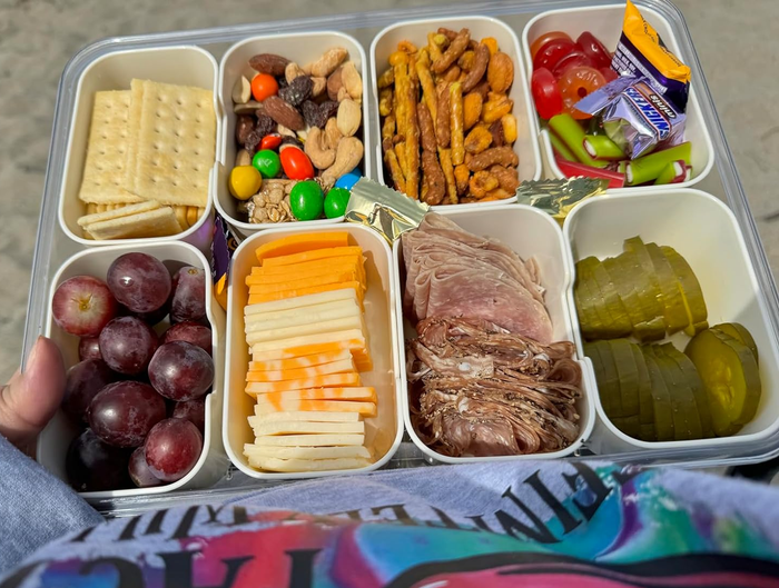 A reviewer holding a snack box with eight compartments, filled with a variety of foods, at the beach.