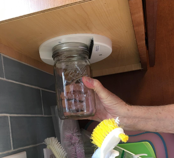 A reviewer’s hand using the jar opener under their kitchen cabinet to twist open a jar
