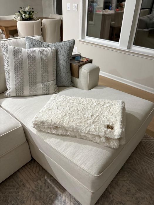A soft, textured white blanket neatly folded on a beige sectional sofa, accented with decorative pillows in a calming, neutral-toned living room.