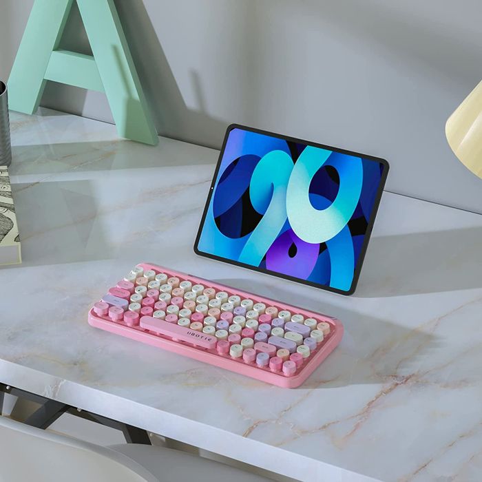 A pink retro-inspired keyboard featuring purple and yellow keys, placed beside a tablet screen.